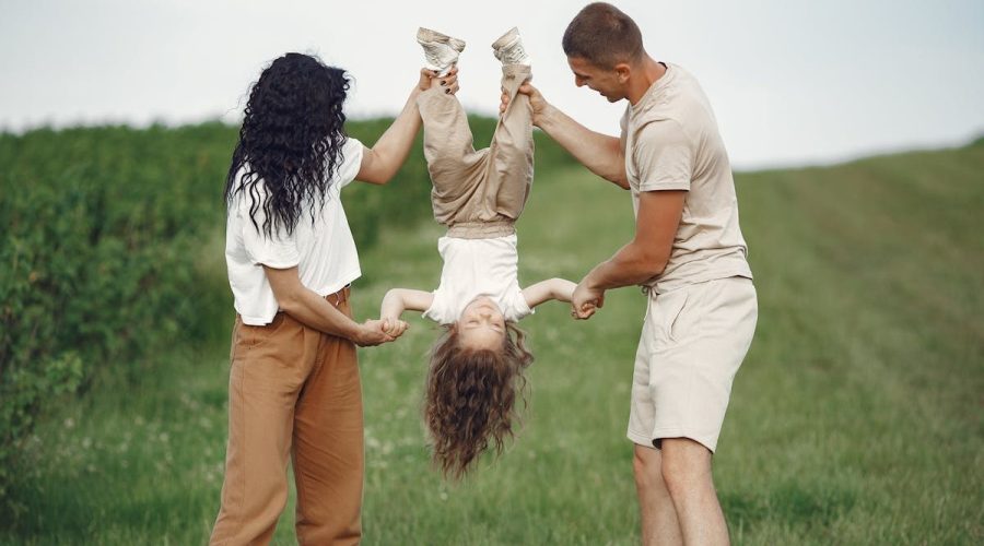 A joyful family enjoys outdoor play, lifting their child upside down in a green field.